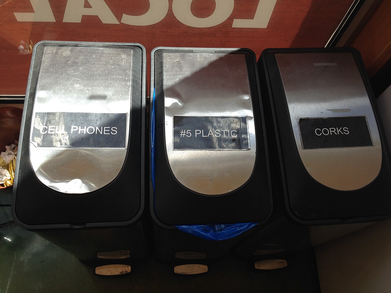 Three black collection bins labeled for cell phones, number-five plastic, and corks stand side by side.
