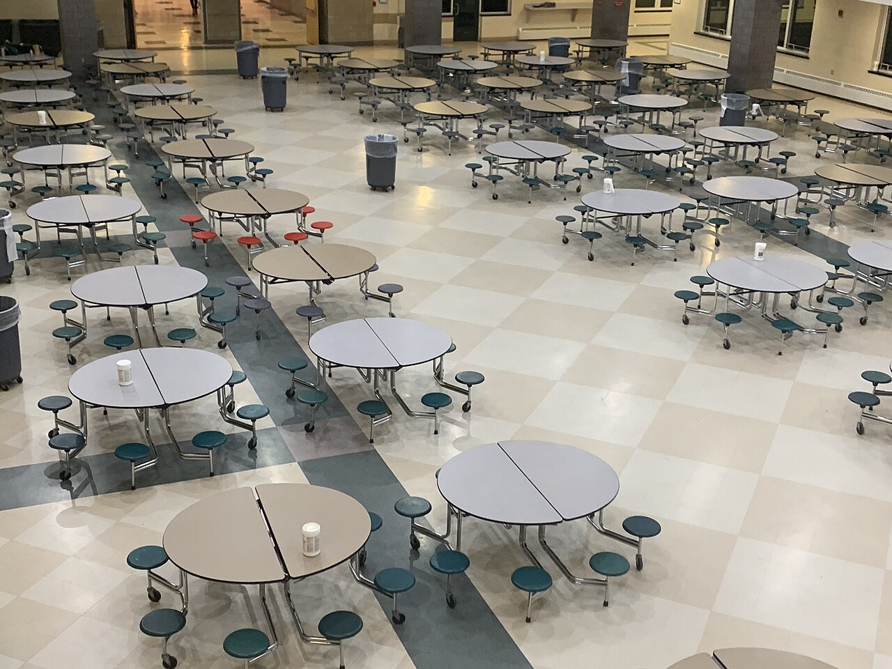 An empty school cafeteria viewed from above, with dozens of circular tables and gray trash cans spaced along the central aisles.